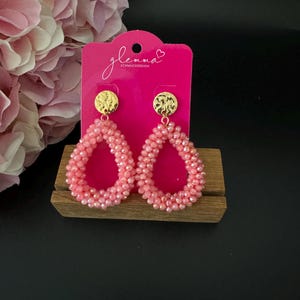 Könnte beinhalten: Ein Paar rosa Perlen-Ohrringe mit goldfarbenen Oberteilen. Die Ohrringe werden auf einer rosa Karte mit dem Text "glemma SCHMUCKDESIGN" präsentiert.