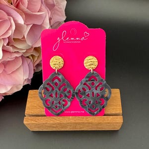 Puede incluir: Un par de pendientes negros y dorados con un diseño geométrico. Los pendientes están en una tarjeta rosa con el texto "glemma SCHMUCKDESIGN" y "www.glemma.me".