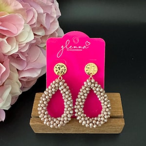 May include: A pair of gold and blush pink teardrop shaped earrings with a hammered gold disc at the top. The earrings are on a pink card with the text "glemma Schmuckdesign".