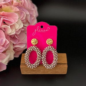 May include: A pair of oval-shaped earrings with a gold-toned metal top and a pink beaded design. The earrings are displayed on a wooden stand with a pink background.