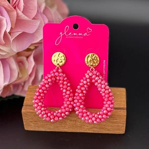 Könnte beinhalten: Hellrosa Perlen-Ohrringe in Tropfenform mit goldfarbenen, kreisförmigen Akzenten. Die Ohrringe werden auf einer pinkfarbenen Karte mit dem Text "glemma SCHMUCKDESIGN" und einer Website-Adresse präsentiert. Auf einer Holzoberfläche.