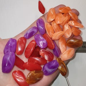 Puede incluir: Una colección de cuentas de forma ovalada en varios colores, incluyendo morado, rojo, naranja y marrón con un acabado jaspeado. Las cuentas se muestran en una palma junto a una regla metálica que muestra una parte de la escala.