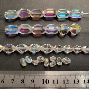 Peut inclure: Plusieurs rangées de perles de cristal iridescentes sont présentées sur un fond sombre. Les perles sont de formes variées, notamment des octogones facettés et des formes irrégulières plus petites. Une règle métallique avec des graduations en centimètres est en bas.