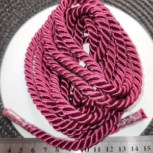 Peut inclure: Une corde enroulée, de couleur bordeaux, avec un design torsadé. La corde est mesurée contre une règle, montrant une longueur d'environ 38 cm. Les extrémités de la corde sont scellées avec du plastique transparent.