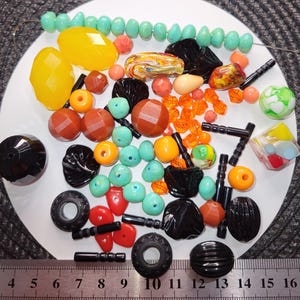 Peut inclure: Une collection de perles colorées de différentes formes et tailles, dont des perles jaunes, oranges et noires facettées, ainsi que des perles rondes turquoise et orange. Une règle est au bas de l'image.