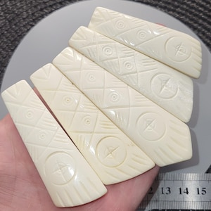 May include: Six ivory-coloured, rectangular decorative pieces are shown. Each piece has carved geometric patterns, including circles, diamonds, and a compass rose design. The pieces are held in a hand, with a ruler visible in the background.