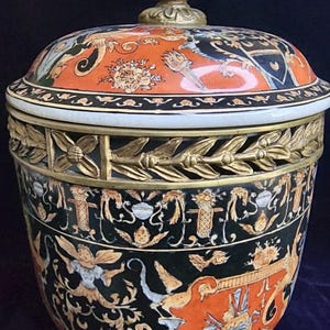 May include: A lidded ceramic jar featuring a black and orange ground with intricate painted motifs of winged figures, floral elements, and heraldic designs. The jar is accented with a gold-toned metal band of laurel leaves and a pineapple-shaped finial.