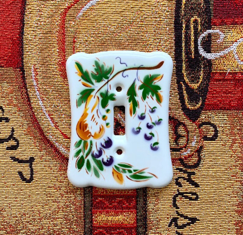 May include: A white ceramic light switch cover with a hand-painted design of grapes and leaves in shades of green, yellow, and purple.