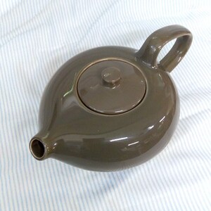 May include: A brown ceramic teapot with a lid. The teapot has a rounded body and a spout on the side. The teapot is sitting on a white and blue striped surface.