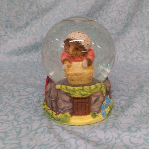 May include: A glass snow globe with a small hedgehog figurine inside. The hedgehog is wearing a pink dress and a white bonnet and is holding a basket of yellow flowers. The base of the snow globe is a brown and gray stone structure with a small door and green vines.