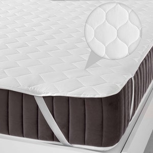 May include: White quilted mattress pad with elastic straps to secure it to a mattress. The pad has a diamond pattern and is shown on a dark gray mattress.
