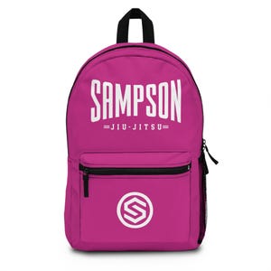 Op de afbeelding: Roze rugzak met een wit logo met de tekst "SAMPSON =JIU-JITSU=". Het logo bevindt zich op de voorkant van de rugzak. Op de voorkant van de rugzak bevindt zich onderaan een witte cirkel met een letter "S" erin.
