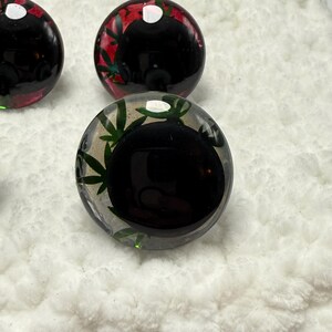 Peut inclure: Gros plan de plusieurs billes de verre rondes et transparentes avec un centre noir. Certaines billes ont des accents verts et rouges avec des motifs de feuilles. Les billes sont sur une surface blanche et textur&eacute;e.