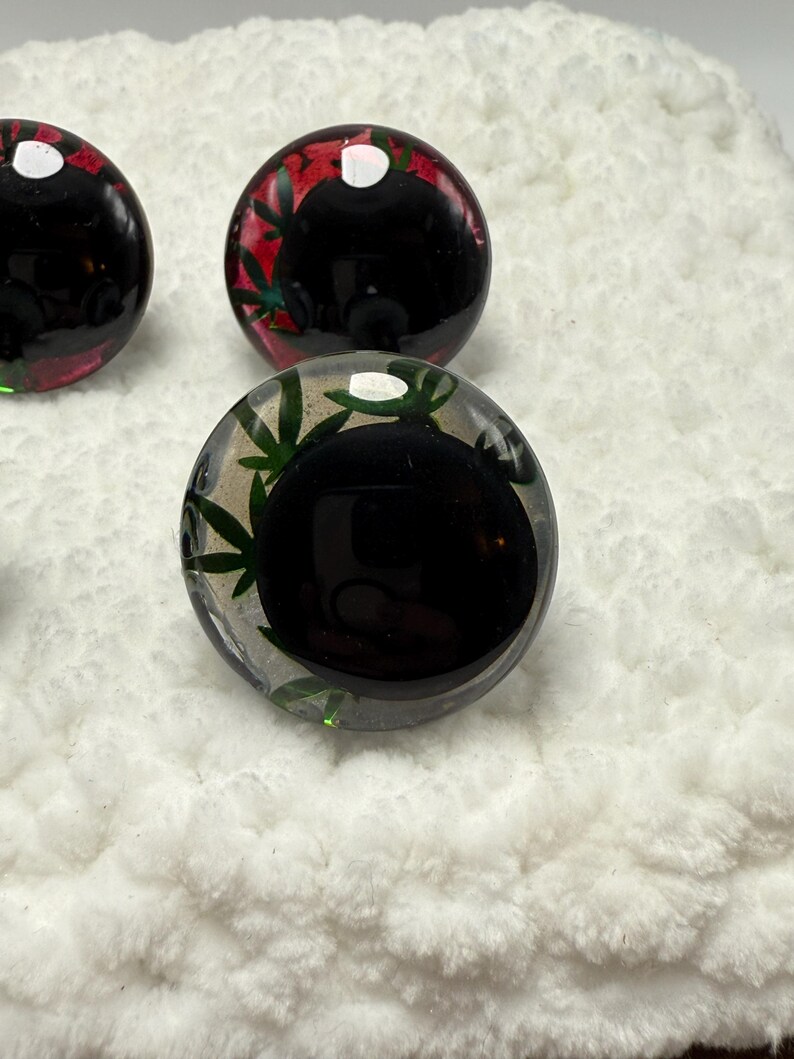 Peut inclure: Gros plan de plusieurs billes de verre rondes et transparentes avec un centre noir. Certaines billes ont des accents verts et rouges avec des motifs de feuilles. Les billes sont sur une surface blanche et textur&eacute;e.