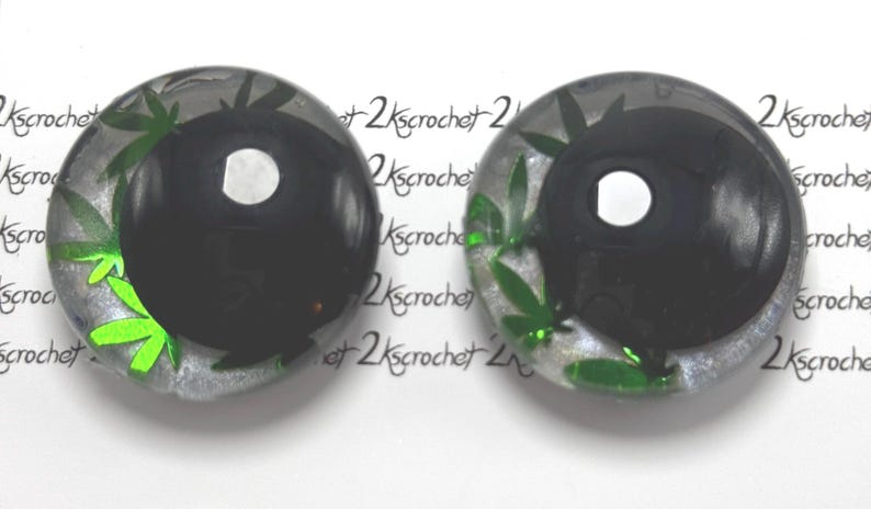Peut inclure: Deux cabochons ronds en r&eacute;sine transparente avec un motif yin-yang noir. Le motif est orn&eacute; de feuilles de cannabis vertes. Le fond est un glitter argent&eacute;. Le texte "2kscrochet" est r&eacute;p&eacute;t&eacute; autour des cabochons.