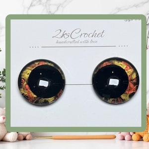 May include: Two round, black doll eyes with gold and red accents, displayed on a white card with the text "2ks Crochet handcrafted with love". The eyes are connected by a thin wire.