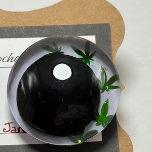 Peut inclure: Un presse-papier rond et transparent avec un motif yin-yang noir et blanc. Des accents verts en forme de feuilles sont dispers&eacute;s sur les bords. Le presse-papier est sur une surface blanche avec le texte "sCrochet" et "rah Jan" visible.