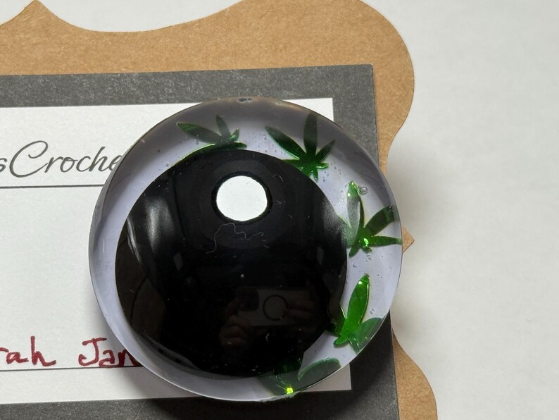 Peut inclure: Un presse-papier rond et transparent avec un motif yin-yang noir et blanc. Des accents verts en forme de feuilles sont dispers&eacute;s sur les bords. Le presse-papier est sur une surface blanche avec le texte "sCrochet" et "rah Jan" visible.