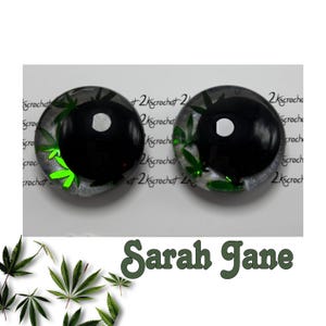 Peut inclure: Deux boucles d'oreilles rondes, noires et transparentes, avec un motif de feuille de cannabis. Les boucles d'oreilles ont un centre noir avec un point blanc et des feuilles de cannabis vertes sur les bords. Le texte "Sarah Jane" est en dessous.