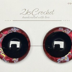 May include: Two round, red and black craft eyes with a glossy finish. The outer ring has a marbled effect with gold accents. The center is a solid black circle. The text "2ks Crochet handcrafted with love" is at the top.