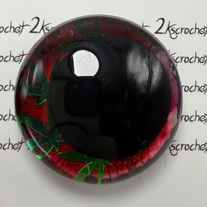 Peut inclure: Cabochon rond et brillant noir avec un motif rouge et vert. Le cabochon a une surface r&eacute;fl&eacute;chissante et pr&eacute;sente des formes vertes en forme de feuilles sur un fond rouge. Le texte "2kscrochet" est &eacute;crit sur le bord du cabochon.