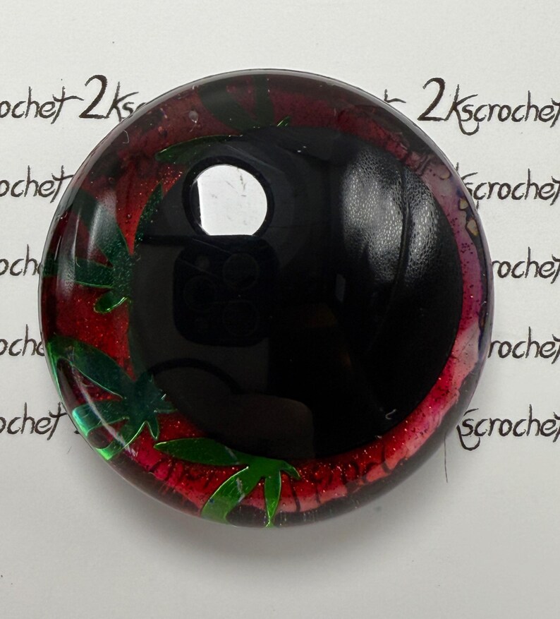Peut inclure: Cabochon rond et brillant noir avec un motif rouge et vert. Le cabochon a une surface r&eacute;fl&eacute;chissante et pr&eacute;sente des formes vertes en forme de feuilles sur un fond rouge. Le texte "2kscrochet" est &eacute;crit sur le bord du cabochon.