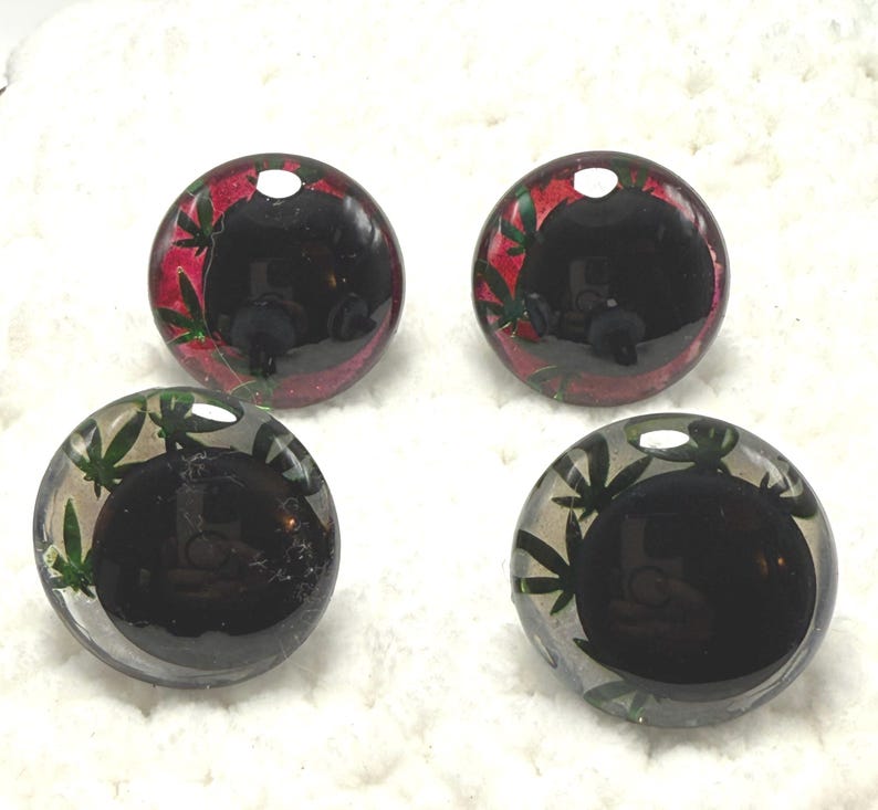 Peut inclure: Quatre cabochons ronds et transparents avec un centre sombre. Deux ont un motif de feuilles rouges et vertes sur le bord, et deux ont un motif de feuilles vertes. Les cabochons sont pos&eacute;s sur un fond blanc textur&eacute;.