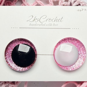 May include: Two round, pink glitter-edged craft eyes with black and white centers, displayed on a white card. The card reads "2ks Crochet handcrafted with love". The background features pink crochet.