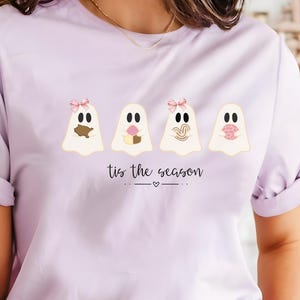 May include: A light purple t-shirt with rolled-up sleeves featuring four ghost illustrations with bows and the text "tis the season". The shirt is a soft, comfortable material.