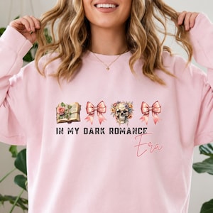 Può includere: Una felpa a maniche lunghe rosa chiaro con una stampa grafica raffigurante un libro aperto, due fiocchi rosa e un teschio floreale. Il testo IN MY DARK ROMANCE Era è scritto in caratteri neri e rosa sul petto del capo di abbigliamento.