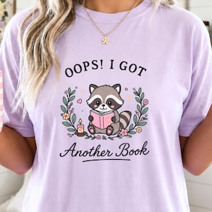 Puede incluir: Una camiseta de color morado claro con un gráfico de un mapache leyendo un libro rosa, rodeado de coronas de flores y una pequeña vela. El texto sobre el mapache dice OOPS! I GOT y el texto debajo dice Another Book en letras negras.