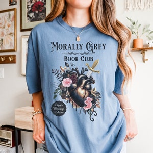 May include: A light blue t-shirt with the text "Morally Grey Book Club." The graphic features a heart pierced by a sword, surrounded by flowers. The shirt also has the text "Trigger Warnings Welcome."