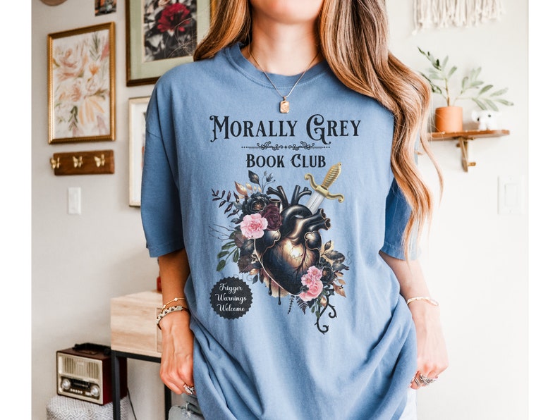 May include: A light blue t-shirt with the text "Morally Grey Book Club." The graphic features a heart pierced by a sword, surrounded by flowers. The shirt also has the text "Trigger Warnings Welcome."