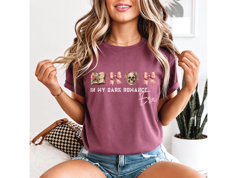 May include: A maroon short-sleeve t-shirt featuring a graphic print of an open book, two pink bows, and a floral skull. The text reads IN MY DARK ROMANCE Era in white and pink script lettering across the center of the chest area.