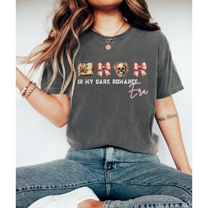 May include: A charcoal gray short-sleeve t-shirt featuring a graphic print with a book, two pink bows, and a floral skull. The text reads IN MY DARK ROMANCE Era in white and pink lettering. The shirt has a crew neckline and a relaxed fit.
