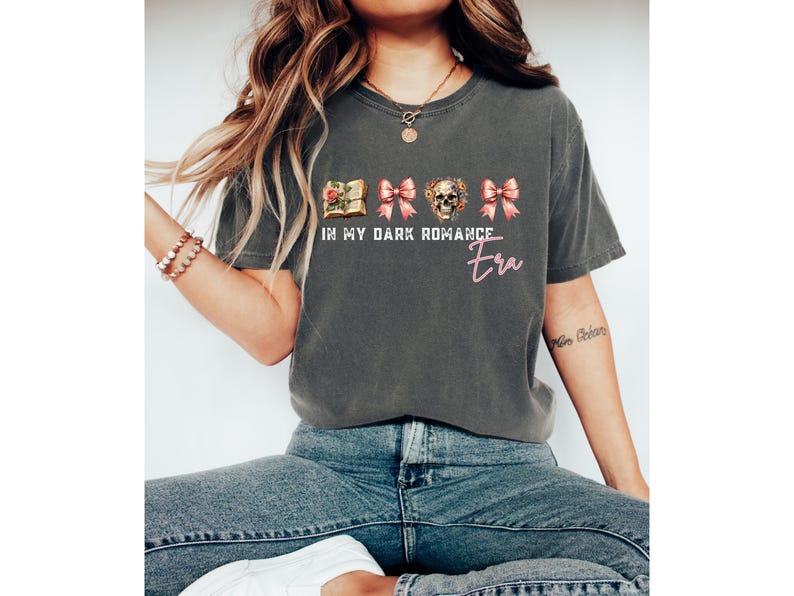 May include: A charcoal gray short-sleeve t-shirt featuring a graphic print with a book, two pink bows, and a floral skull. The text reads IN MY DARK ROMANCE Era in white and pink lettering. The shirt has a crew neckline and a relaxed fit.