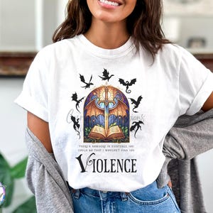 May include: White t-shirt featuring a fantasy-themed graphic. The design includes a sword, open book, dragons, and the word "VIOLENCE". The shirt also has the text "THERE'S NOWHERE IN EXISTENCE YOU COULD GO THAT I WOULDN'T FIND YOU".