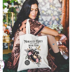 May include: A white tote bag with the text "Morally Grey Book Club" and a graphic of a heart, flowers, and a sword. The bag is held by a person wearing a patterned dress.