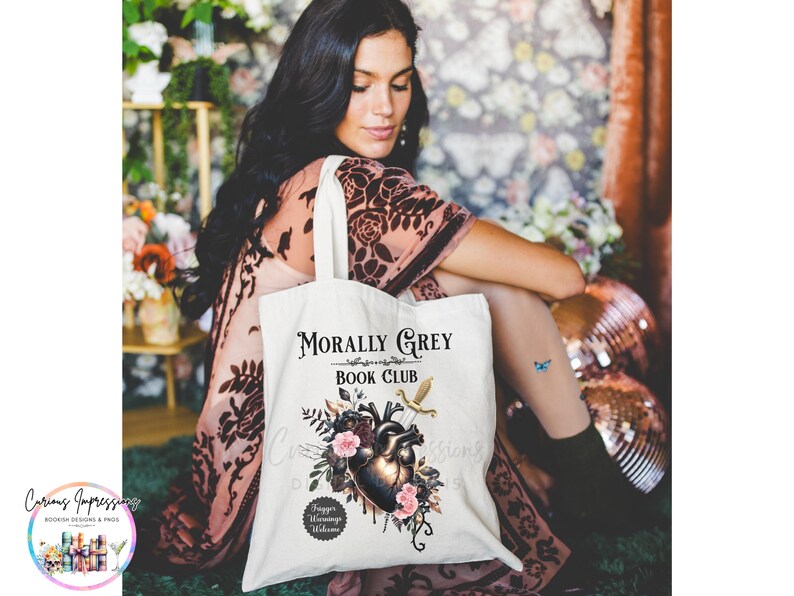 May include: A white tote bag with the text "Morally Grey Book Club" and a graphic of a heart, flowers, and a sword. The bag is held by a person wearing a patterned dress.