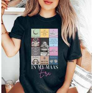 K&ouml;nnte beinhalten: Schwarzes T-Shirt mit einem farbenfrohen Grafikdesign mit Buchcovern und dem Text "IN MY MAAS Era". Das Design enth&auml;lt den Text "BATTLE THE STARS" und "DON'T LET THE HARD DAYS WIN".