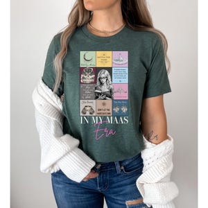 K&ouml;nnte beinhalten: Dunkelgr&uuml;nes T-Shirt mit einer farbenfrohen Grafik mit Buchmotiven und Text. Der Text "IN MY MAAS era" ist in rosa Schrift geschrieben. Das Shirt ist ein l&auml;ssiger Stil.