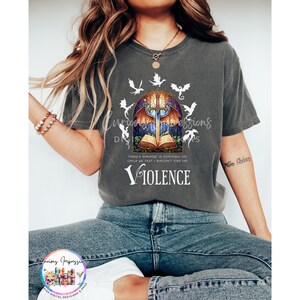 May include: Dark gray t-shirt featuring a stained-glass design with a sword, open book, and dragons. The word "Violence" is printed below the graphic. The shirt has a relaxed fit.