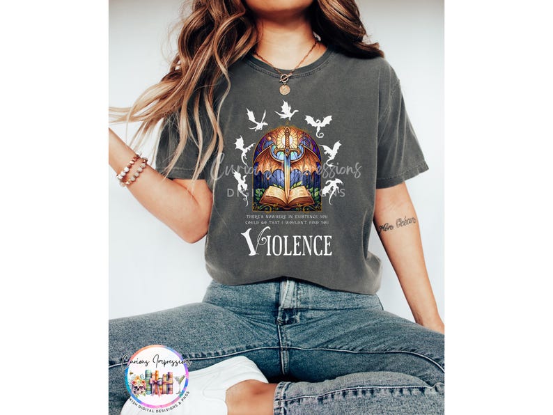 May include: Dark gray t-shirt featuring a stained-glass design with a sword, open book, and dragons. The word "Violence" is printed below the graphic. The shirt has a relaxed fit.