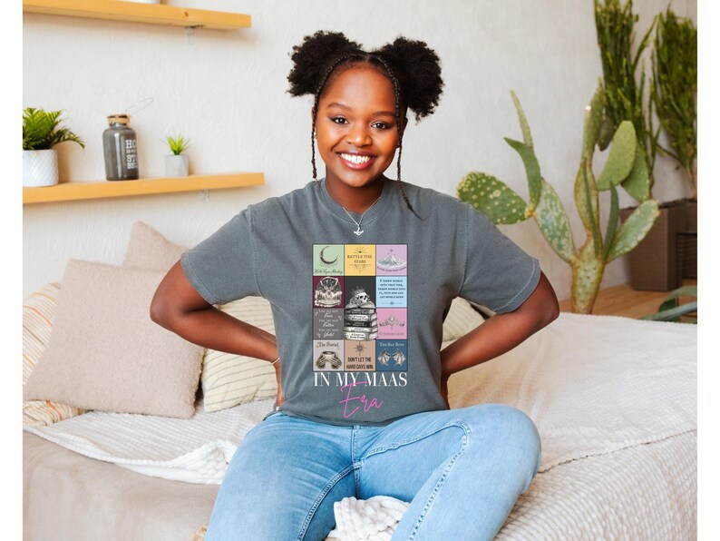 K&ouml;nnte beinhalten: Ein graues T-Shirt mit einem farbenfrohen Grafikdesign mit verschiedenen Bildern und dem Text "IN MY MAAS era" in Pink. Das T-Shirt wird von einer Person getragen.