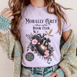 May include: A light purple t-shirt with the text "Morally Grey Book Club" above a graphic of a heart pierced by a sword, surrounded by flowers. The shirt also has a small graphic that says "Trigger Warnings Welcome".