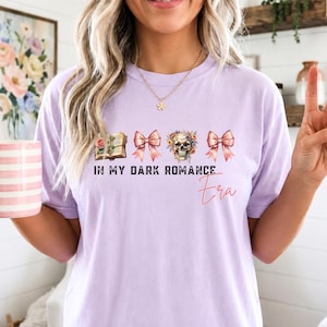 May include: A light purple short-sleeve t-shirt featuring a graphic print of an open book, two pink bows, and a floral skull. The text reads IN MY DARK ROMANCE Era in a combination of bold black block letters and a thin red cursive script.