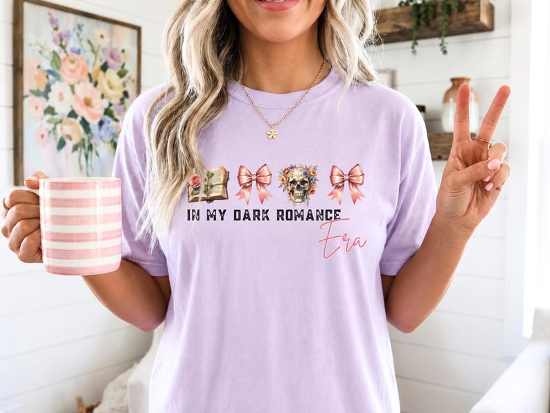May include: A light purple short-sleeve t-shirt featuring a graphic print of an open book, two pink bows, and a floral skull. The text reads IN MY DARK ROMANCE Era in a combination of bold black block letters and a thin red cursive script.