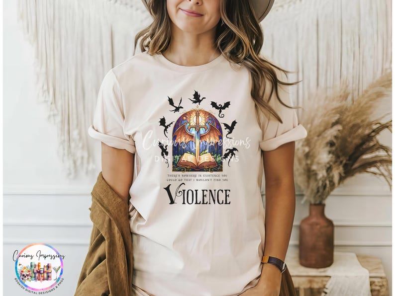 May include: A beige t-shirt featuring a graphic with a sword, open book, and dragons. The text includes "Curious Impressions" and the word "Violence". The shirt has rolled-up sleeves.