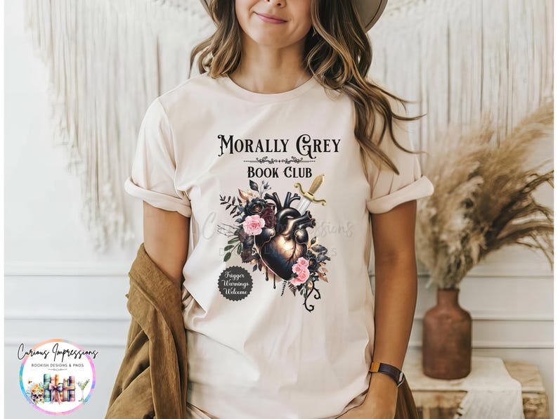May include: Beige t-shirt with the text "Morally Grey Book Club" above a graphic of a heart, flowers, and a sword. The shirt also has the text "Trigger Warnings Welcome".