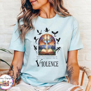 May include: Light blue t-shirt featuring a fantasy-themed graphic. The design includes a sword, open book, stained glass window, and dragons. The word "VIOLENCE" is printed below the graphic. The shirt has short sleeves.